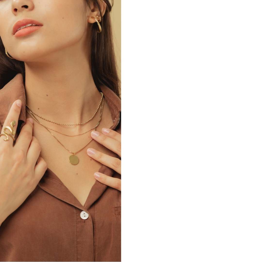 A close-up lifestyle shot of a model wearing layered gold necklaces, including a delicate chain necklace with a round disc pendant and a thicker chain necklace, along with a gold ring on her finger.