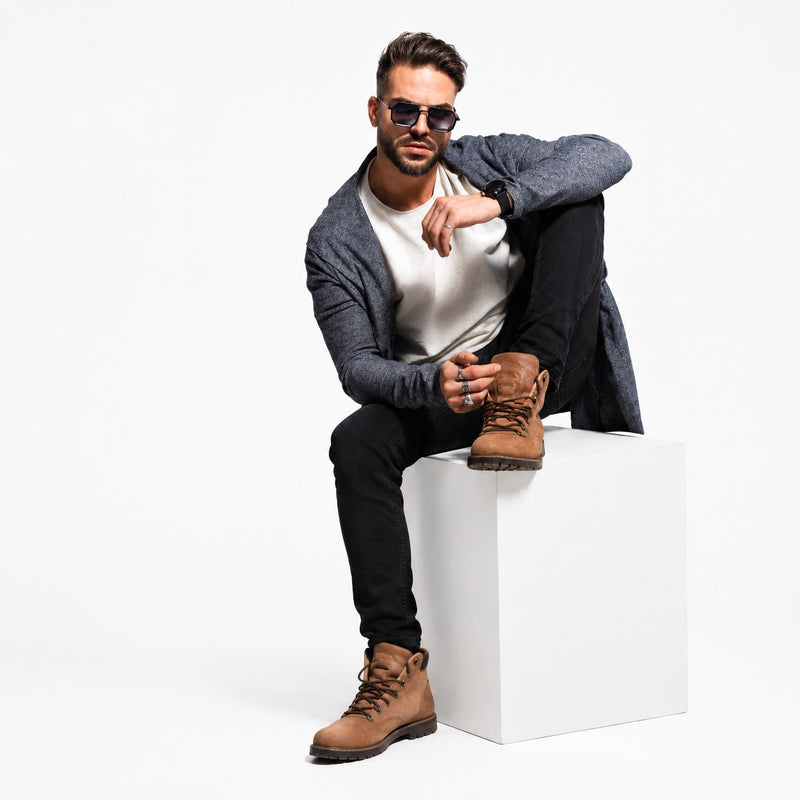 A man wearing sunglasses, a gray cardigan, a white t-shirt, black pants, and brown lace-up boots sits on a white cube against a white studio background.