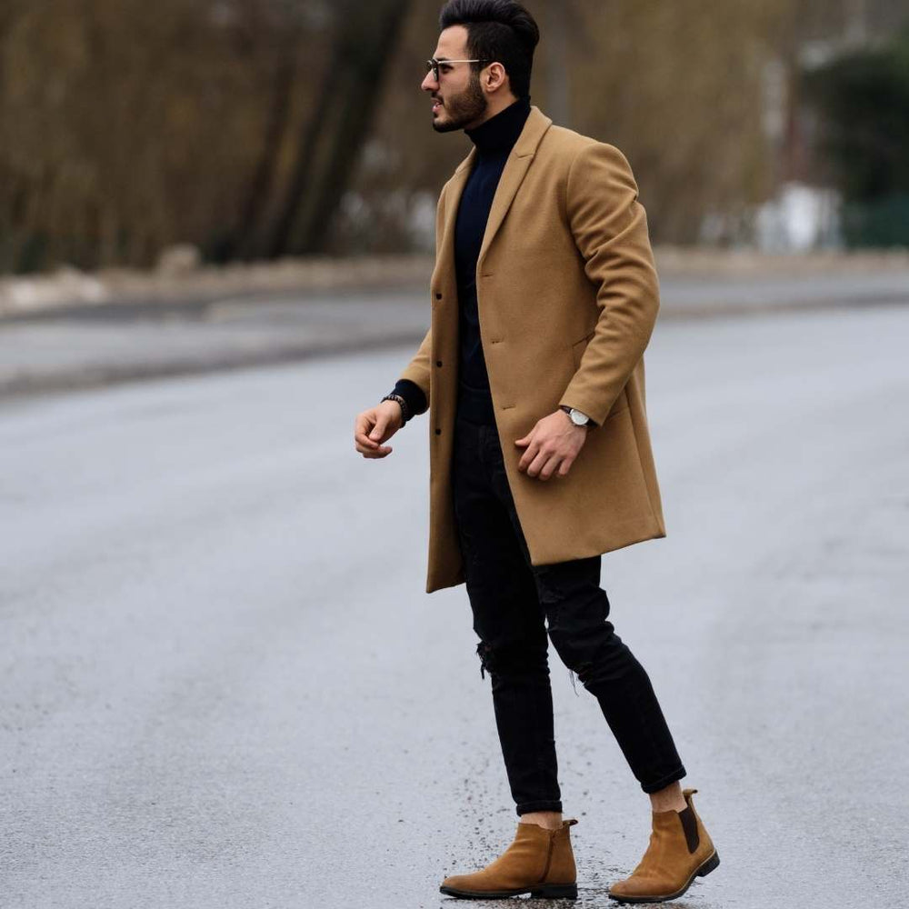 A man walking outdoors on a wet road wearing a camel-colored overcoat, black turtleneck, black jeans, and tan ankle boots.