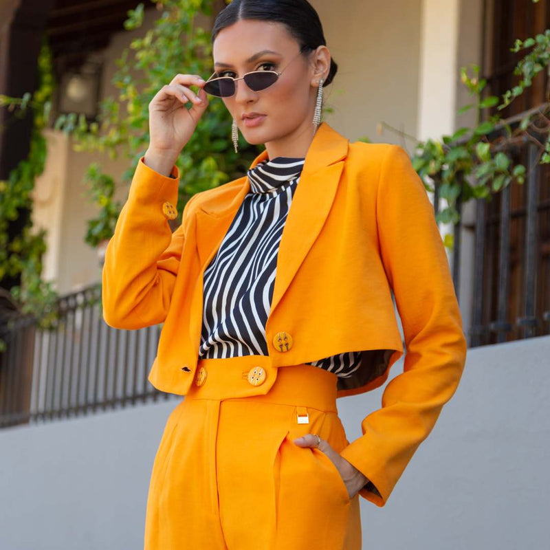 A woman wearing a bright orange tailored blazer and matching trousers over a black-and-white striped top, with small gold buttons and gold-toned sunglasses, photographed outdoors in front of a building with greenery.