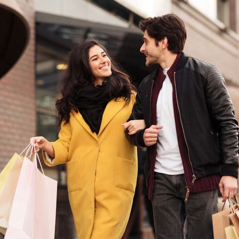 A smiling woman in a mustard yellow coat and black scarf walks arm in arm with a man wearing a black leather-shoulder jacket, white shirt, and dark jeans, while holding shopping bags on a city street.
